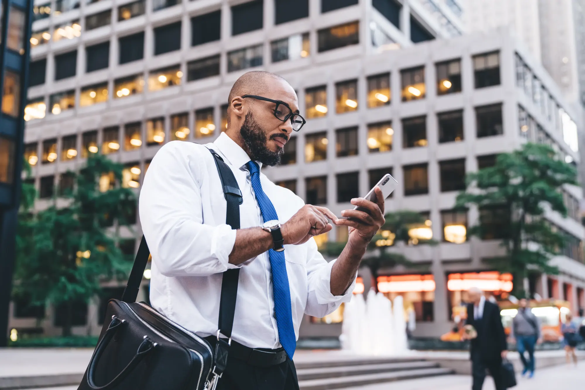 A focused man in a white shirt and blue tie stands in a downtown streetscape, checking his smartphone against the backdrop of large office buildings.