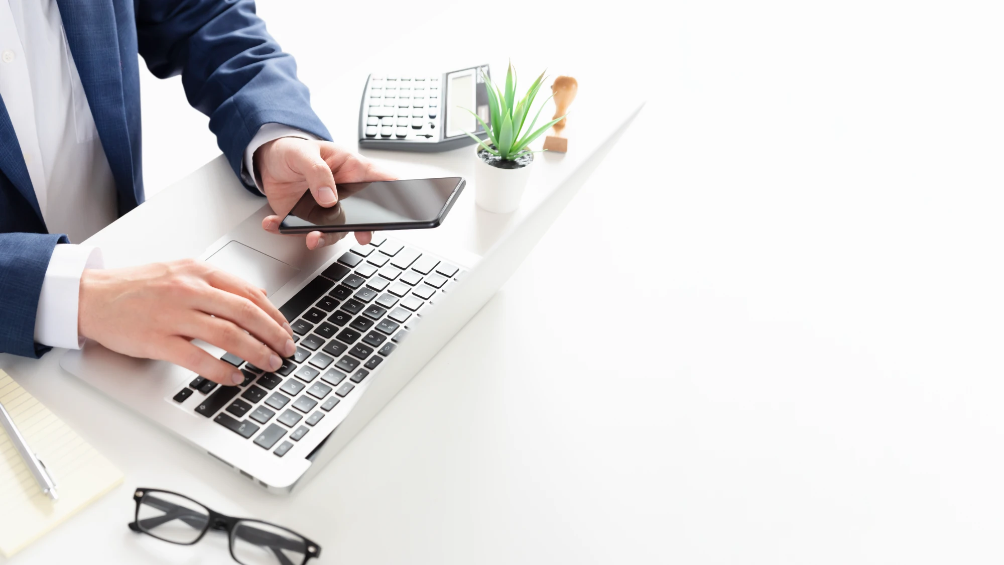 A person in a blue business suit is actively typing on a laptop keyboard with their left hand, while holding a black smartphone in their right hand on a bright white desk.