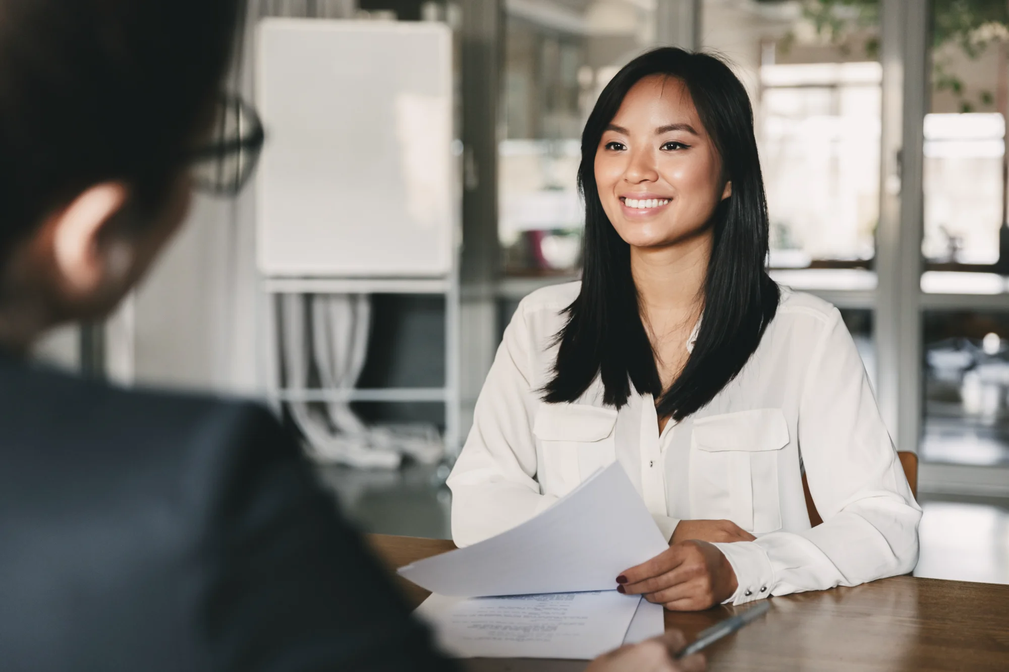 A smiling female job candidate in a white blouse holds papers while attentively looking at the interviewer across the desk in a bright, modern office.
