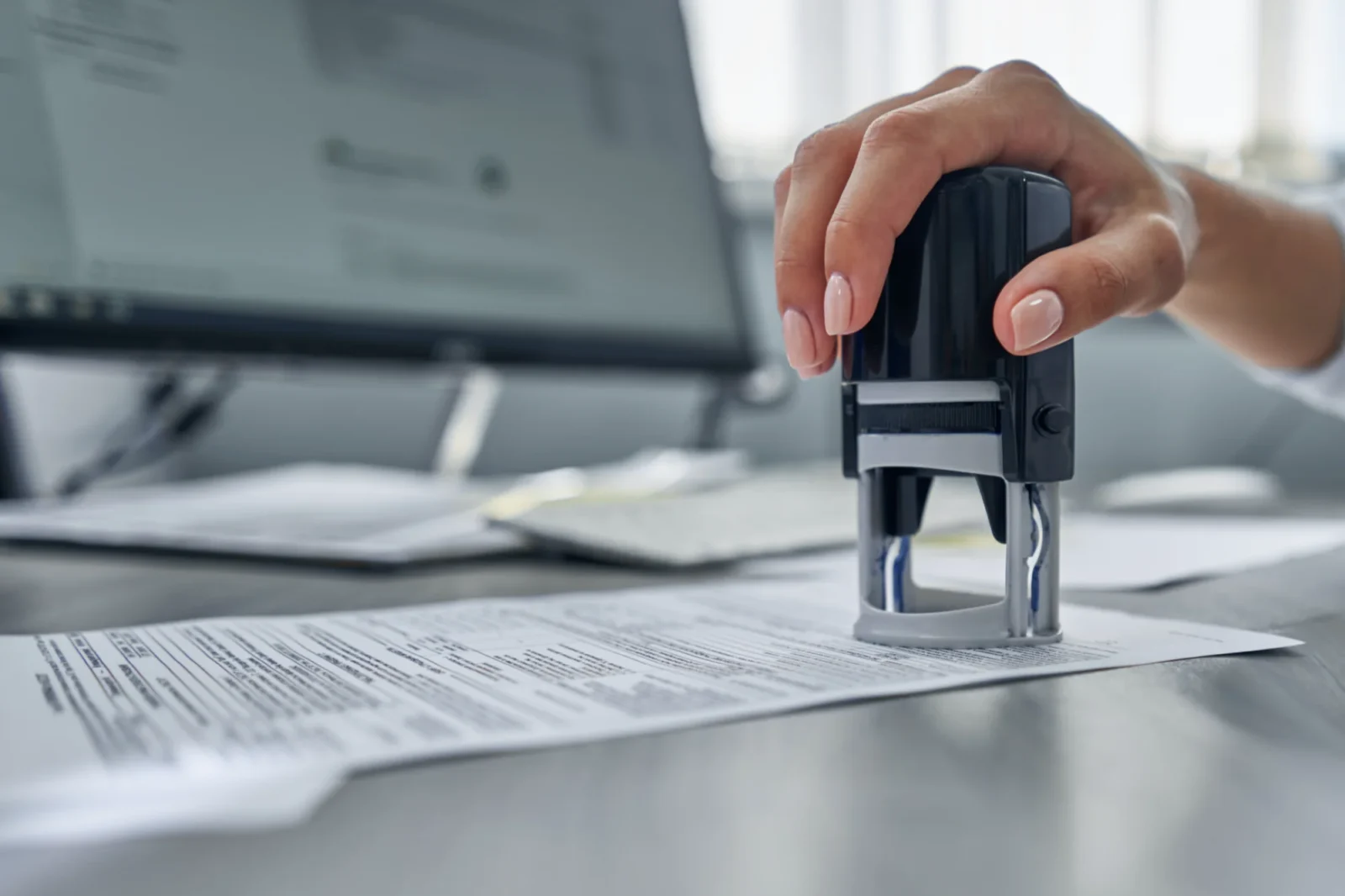 A person's hand with light pink nails holds a black self-inking stamp, poised to press it onto a business document on an office desk.