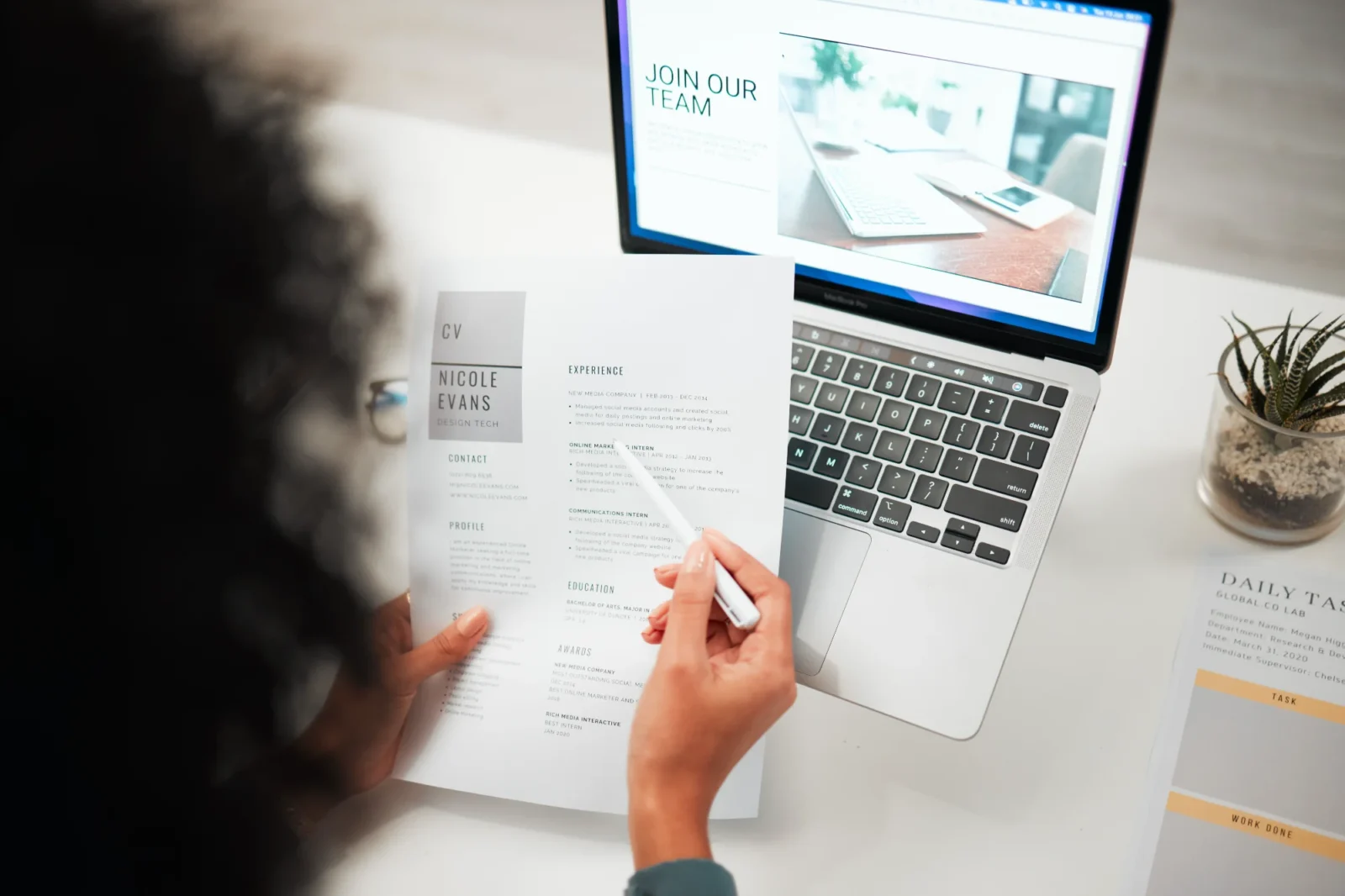 A person at a desk reviews a candidate's resume, pointing to the experience section with a pen. A laptop screen displaying a 'Join Our Team' page is visible.