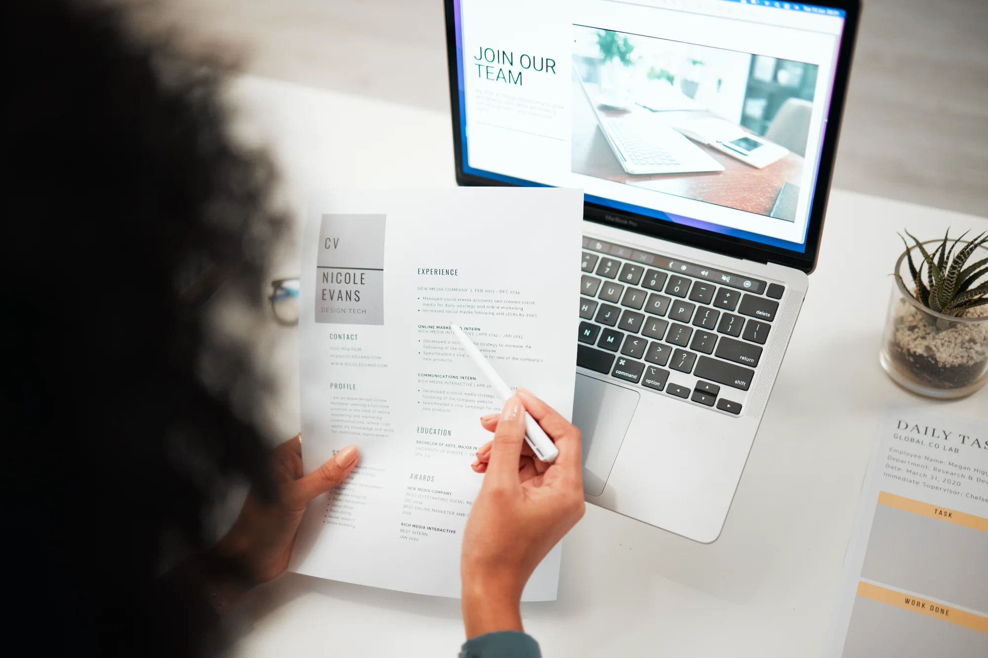 A person at a desk reviews a candidate's resume, pointing to the experience section with a pen. A laptop screen displaying a 'Join Our Team' page is visible.