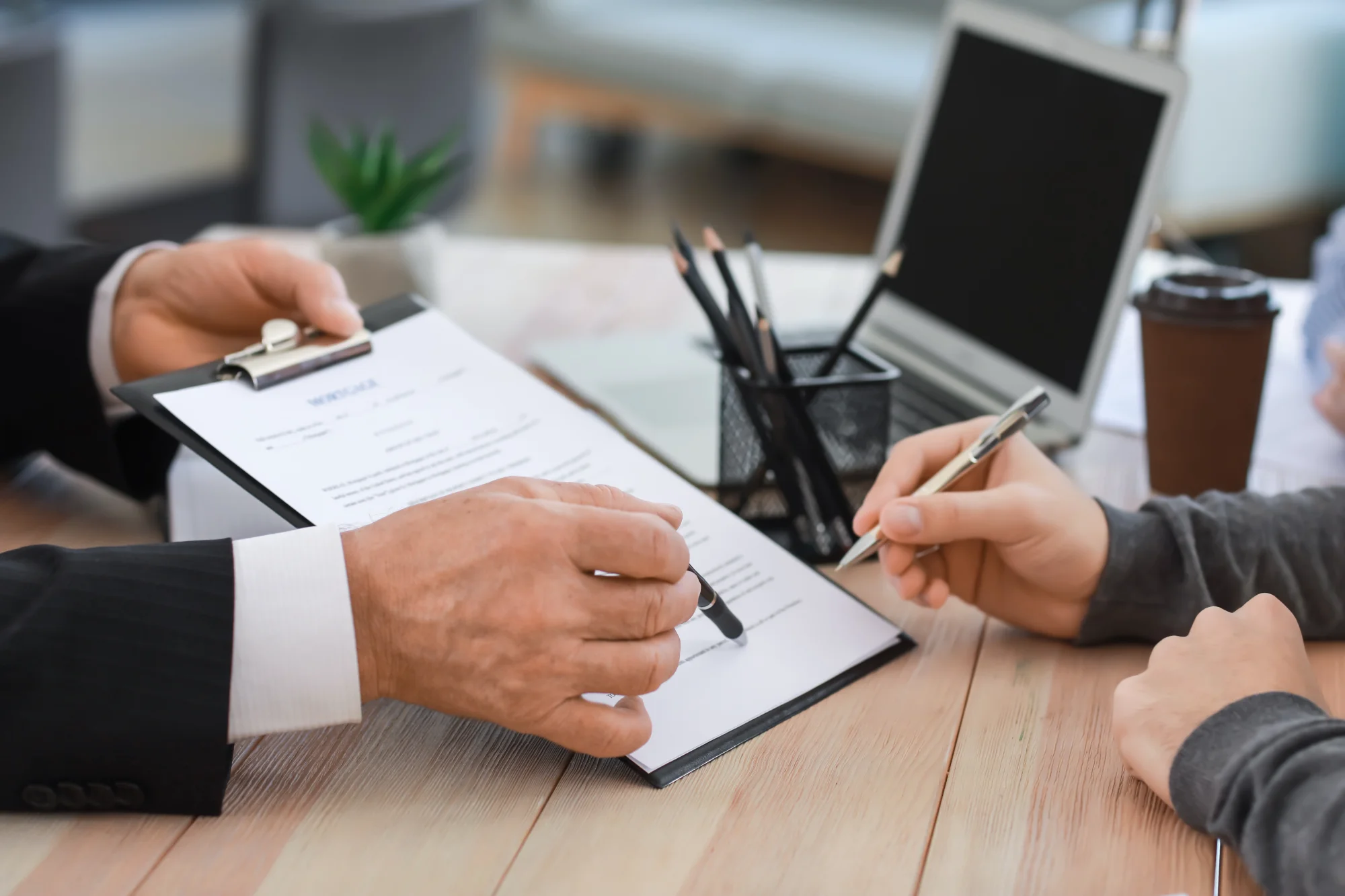 An advisor points with a pen to a contract on a clipboard as a client prepares to sign on a wooden desk near a laptop.