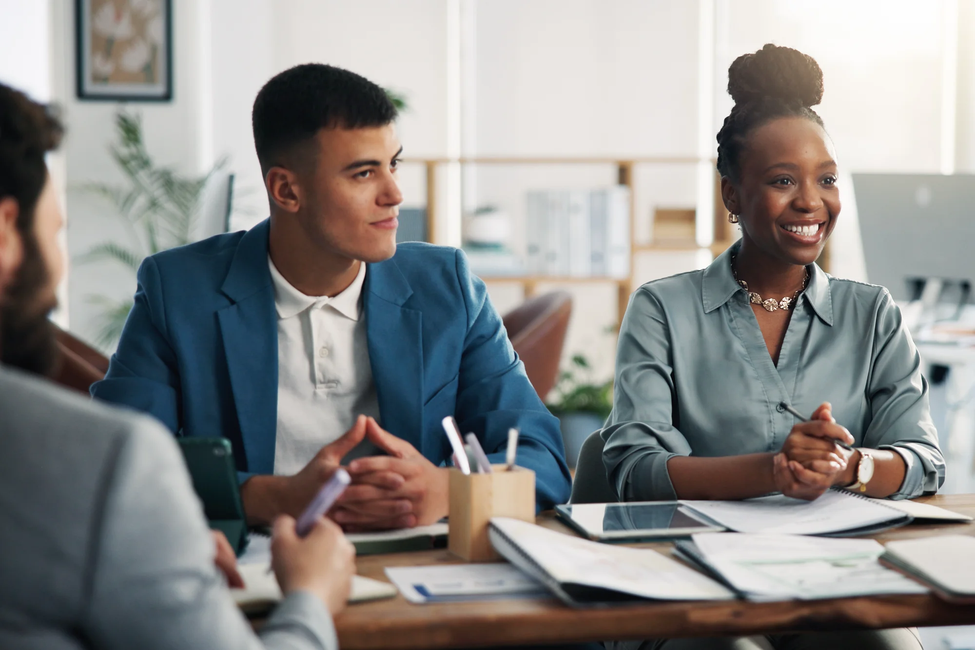 During an office meeting, a smiling woman in a grey blouse and a man in a blue suit sit at a conference table, attentively watching a speaker.