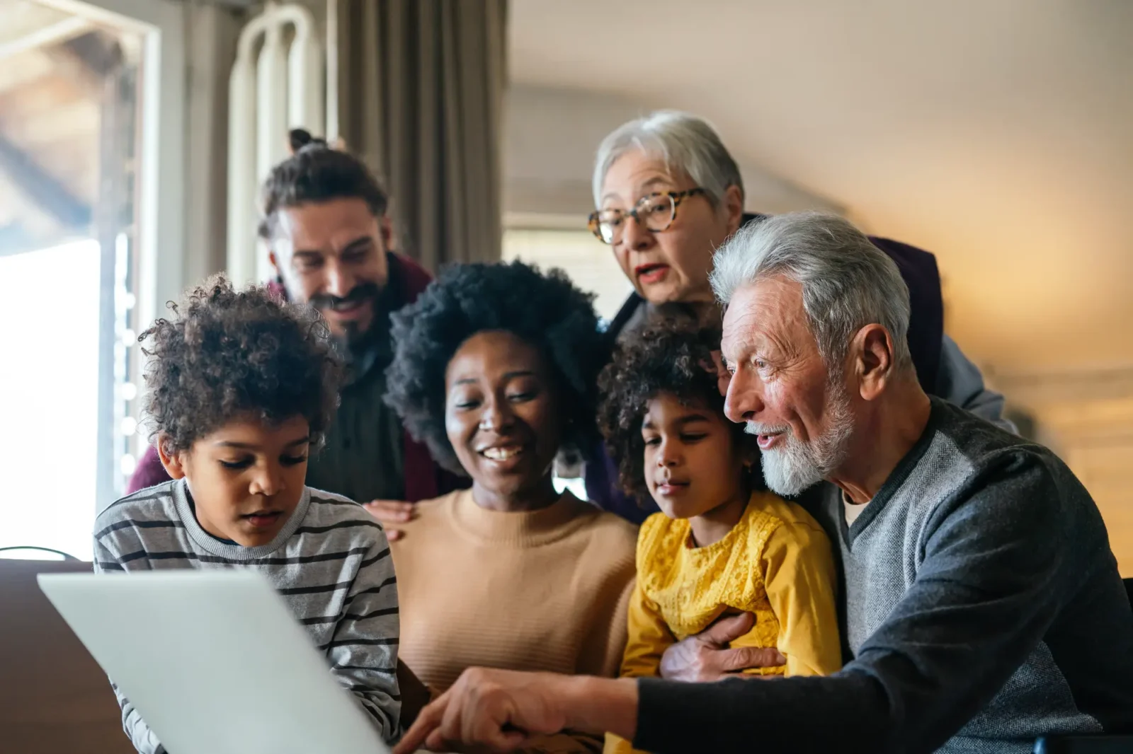 A multi-generational family of six, including grandparents and children, look closely at a laptop screen. The older man points, sharing the screen with the smiling younger adults.