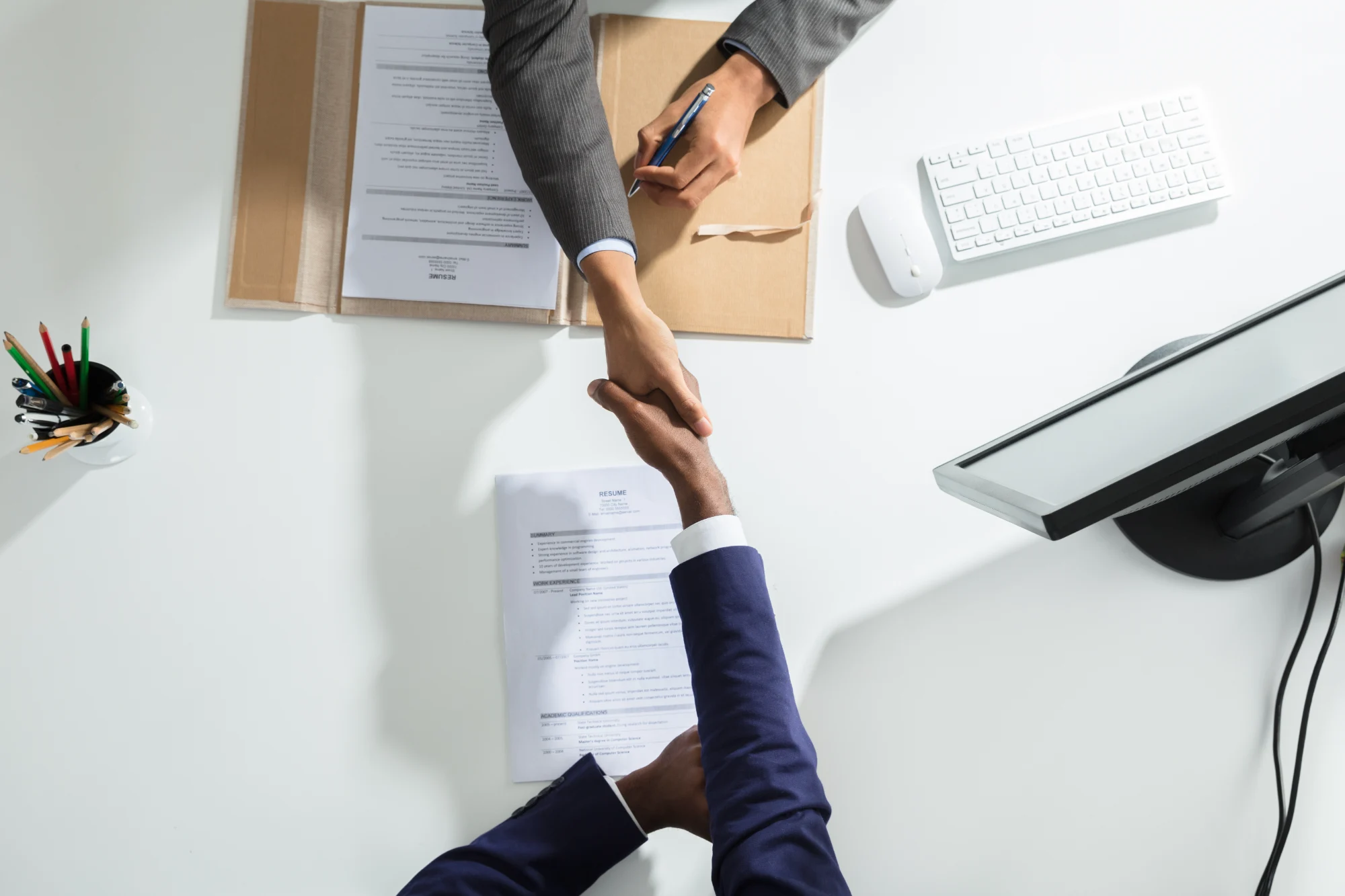An overhead view of two professionals shaking hands across a white desk, with a resume, papers, and office equipment visible nearby.
