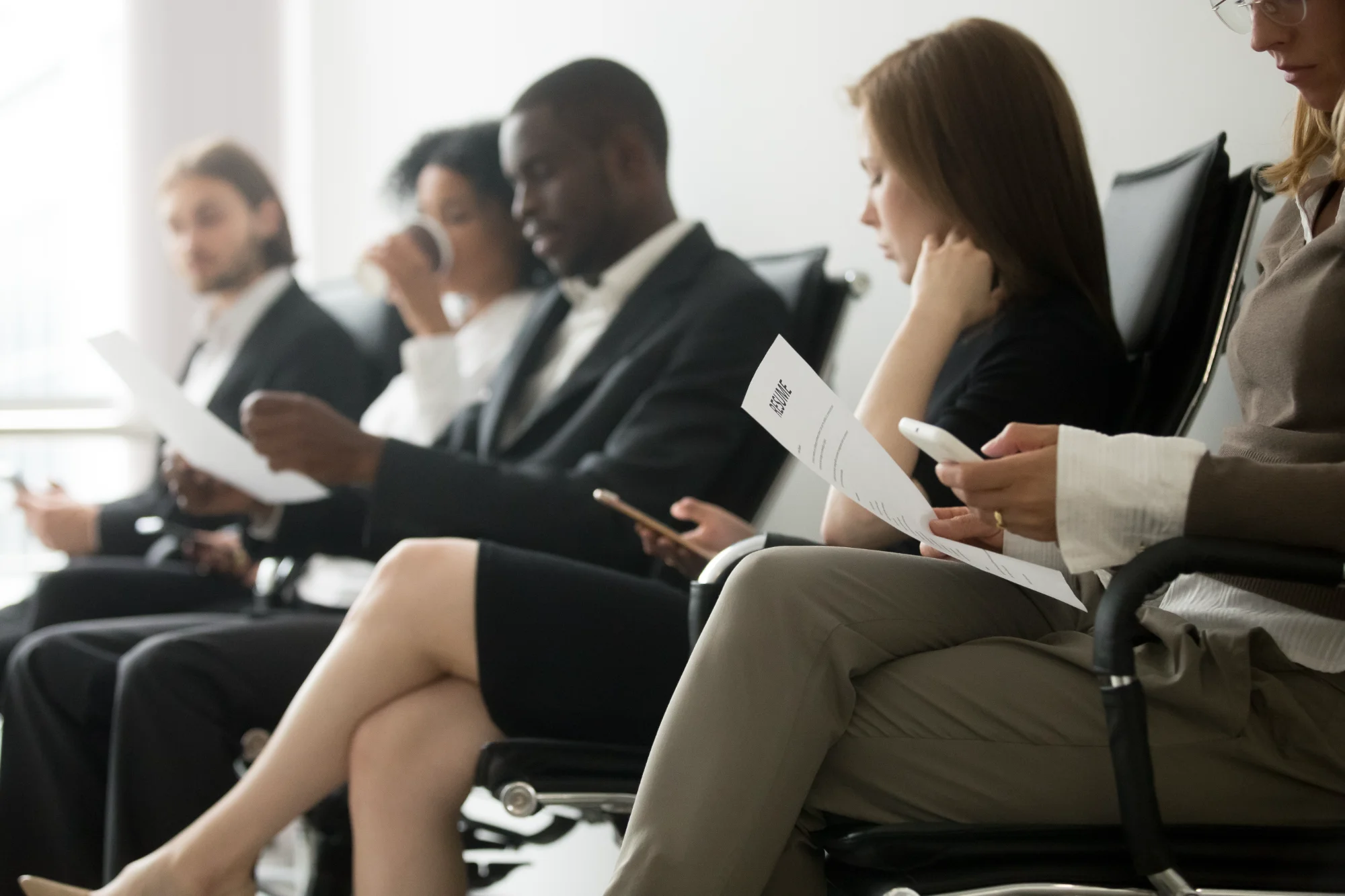 Several professional job candidates in business attire sit attentively in a bright waiting area holding resumes and looking focused.