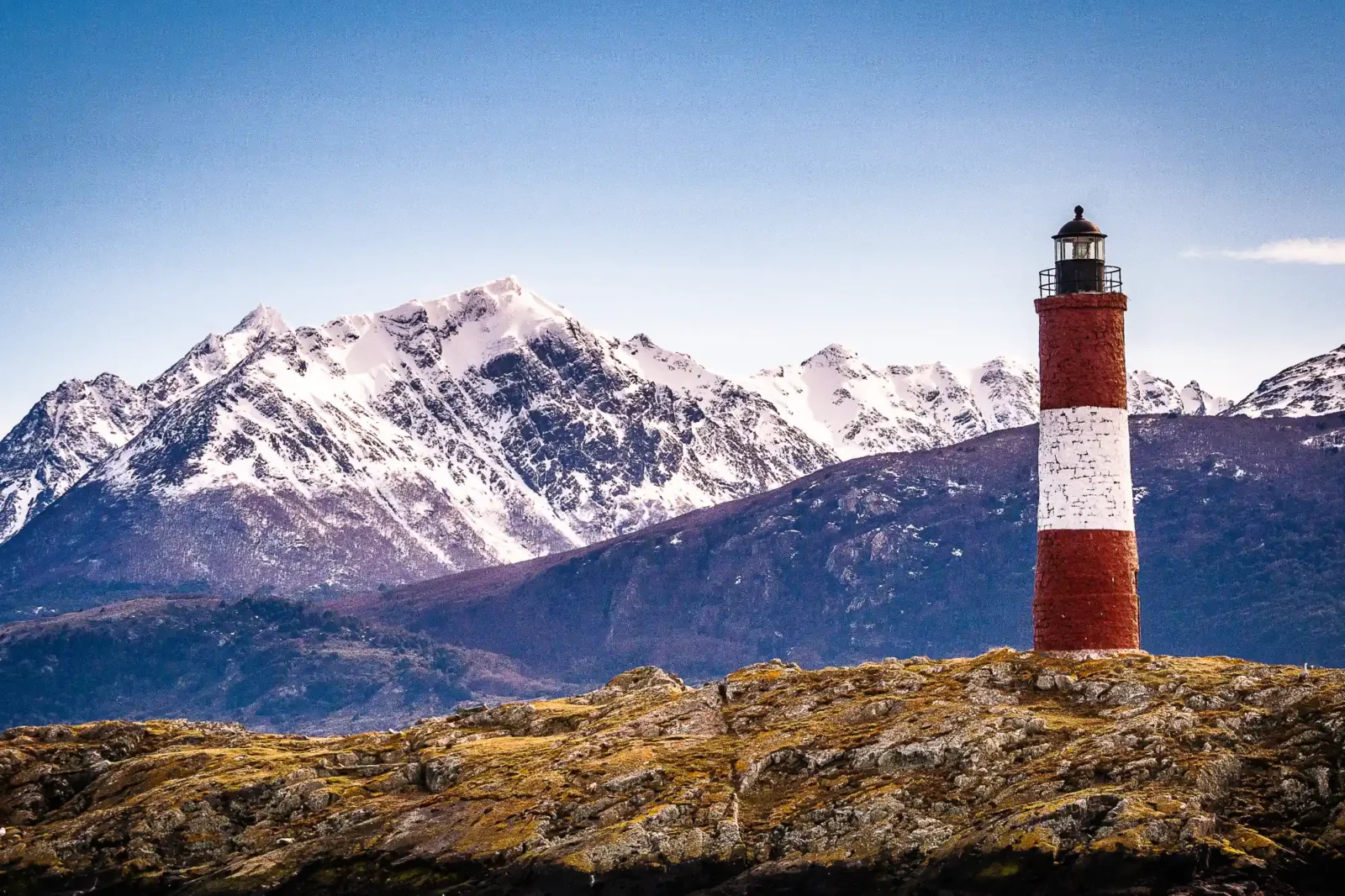 A red and white striped lighthouse stands on a rocky island. Massive snow-covered mountains rise steeply in the background under a clear blue sky.