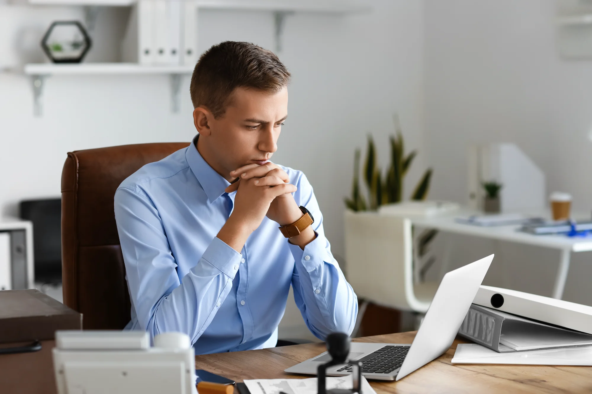 A young notary public in a blue shirt leans forward at his office desk, intently reviewing information on his open laptop screen.