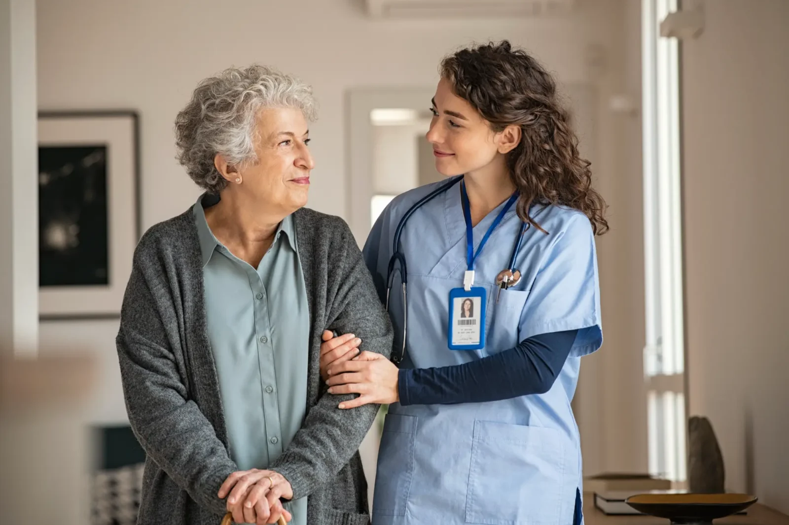 A nurse in blue scrubs supports a senior woman with curly gray hair, who is holding a cane. They are walking indoors and looking at each other with peaceful expressions.