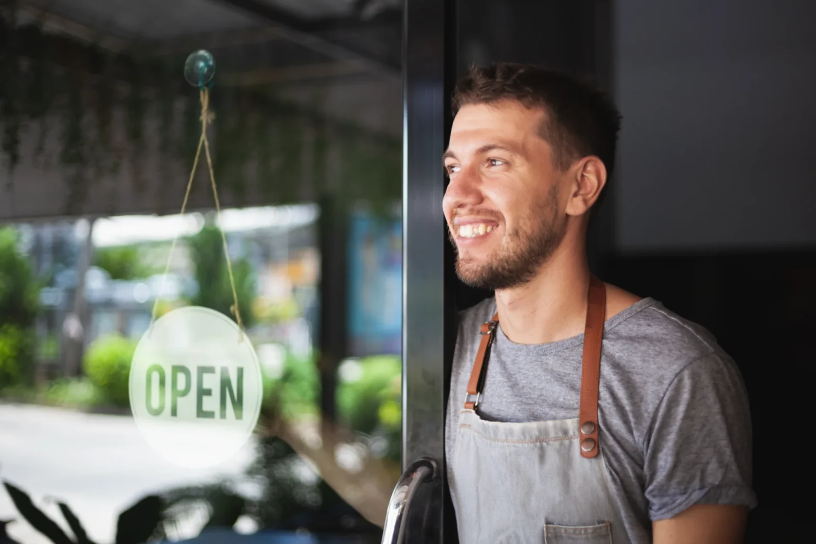 A smiling small business owner in an apron looks outward from his store door, next to a clear glass sign reading "OPEN."