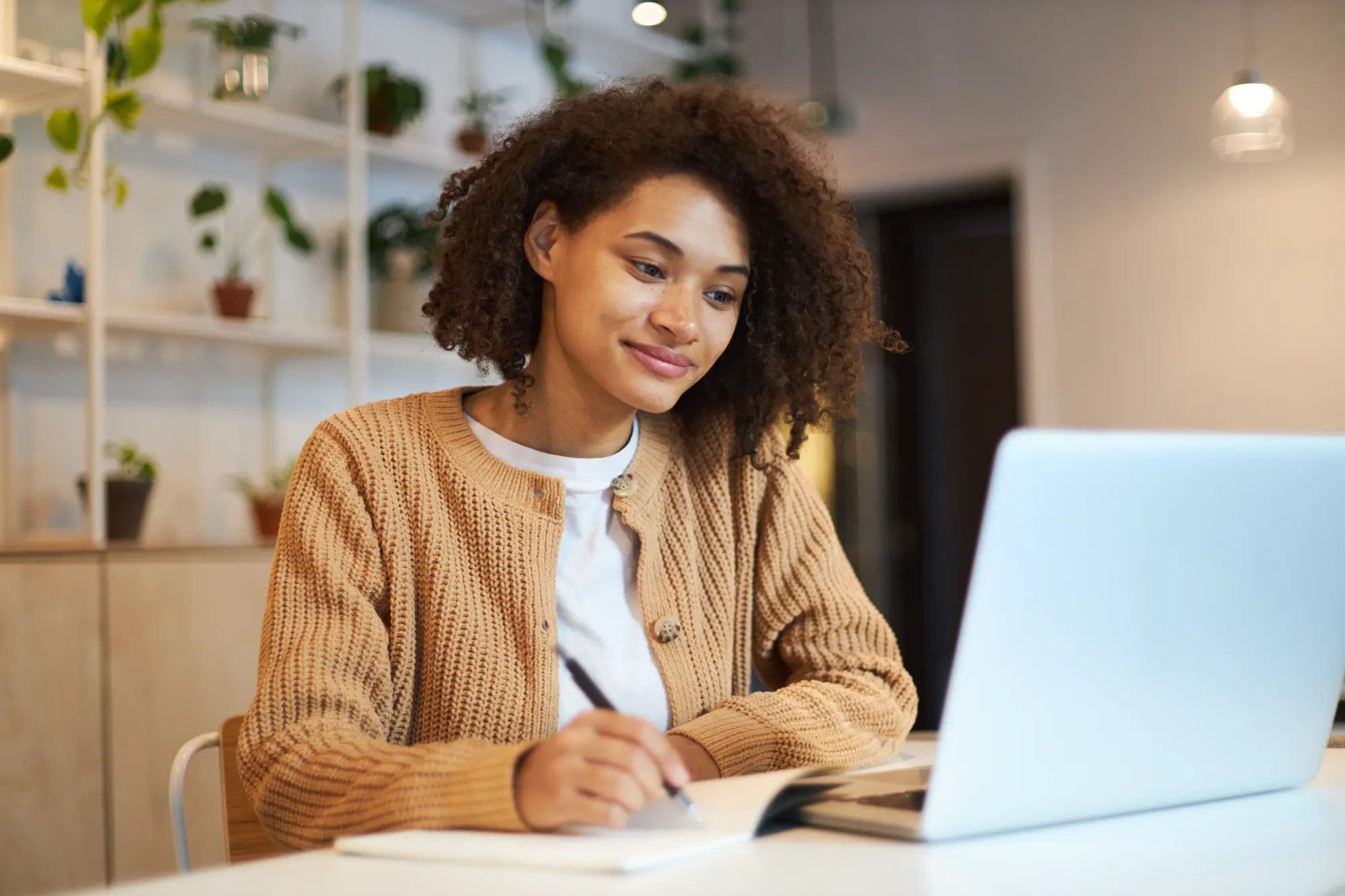A smiling woman in a tan cardigan is taking notes in a notebook while viewing her laptop screen. She is working at a white desk in a bright office environment.